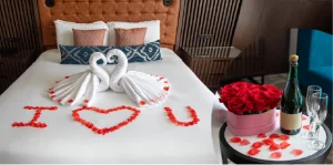 Romantic hotel bed with towel-swans, rose petals spelling 'I love you' on the duvet; side table with a bouquet of roses, champagne and two glasses.