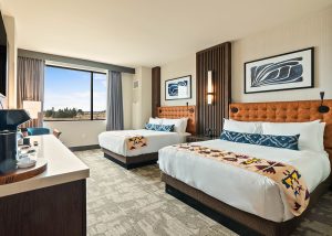 Hotel room with two beds featuring orange tufted headboards, decorative runners, framed abstract art, a large window with curtains, and a coffee station.