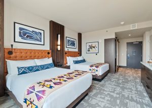 Two queen beds with tufted orange headboards, blue pillows, and colorful geometric runners in a modern hotel room