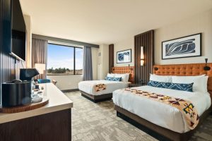 Hotel room with two queen beds, orange tufted headboards, wall art, and a large window with a desk and coffee setup.