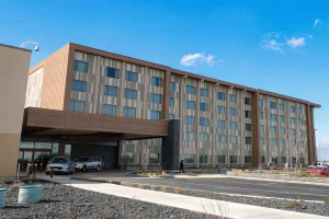 Multi-story hotel exterior with a covered entrance, row of windows, and parked cars in front against a clear blue sky.