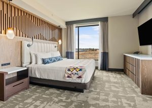 Hotel room with king bed, padded headboard, two wall lamps, and a large window showing a landscape outside.