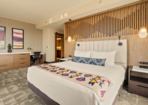 Hotel room with a large bed, padded headboard, and geometric throw; desk area to the left and a doorway to a bathroom in the back-right corner.