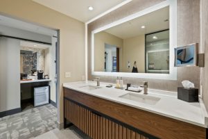 Hotel bathroom vanity with two rectangular sinks, backlit framed mirror, and wood slat cabinet under a white countertop; tissue box on the right.