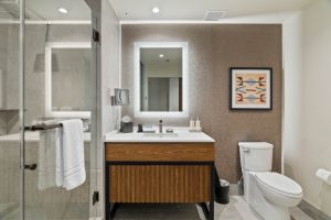 Modern bathroom with a glass-enclosed shower, wood vanity, and a backlit square mirror above a white sink. A framed geometric art piece hangs on a brown accent wall near a toilet.