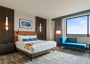 Hotel room with a king bed, tufted headboard, and a bright blue chaise by a large window overlooking a landscape.