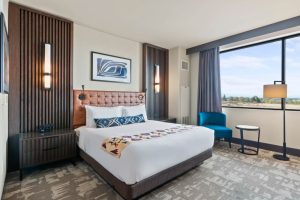 Hotel room with a king bed, tufted brown headboard, two bedside tables, wall art, and warm lamps flanking the bed by a large window.
