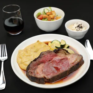 Close-up of a plate with sliced roast beef over mashed potatoes and vegetables, with a glass of red wine in the blurred background.