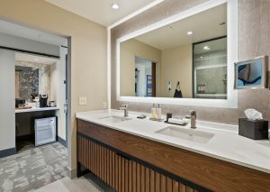 Hotel bathroom with double sinks, a large backlit mirror, and a tissue box; doorway leads to a coffee area and minibar on the left.