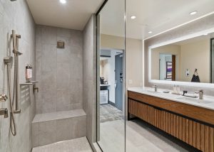 Walk-in shower with built-in bench and multiple metal shower fixtures, glass partition, and gray tile walls next to a double-sink vanity with a backlit mirror.