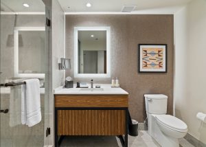 Modern bathroom with glass-enclosed shower, a white countertop sink on a wooden vanity, backlit frameless mirror, and a brown textured accent wall with framed geometric art; toilet and small trash can nearby.