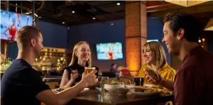 Four friends sit around a table in a lively restaurant, raising drinks and smiling at each other while toasting