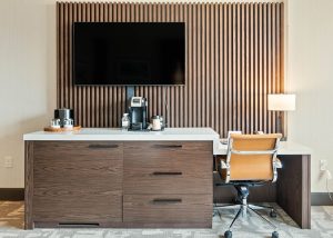 Hotel room workspace with wall-mounted TV above a long wooden cabinet and coffee station on white countertop.