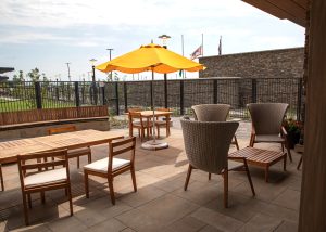 Outdoor patio dining area with wooden tables, cushioned chairs, and a bright yellow umbrella under a clear sky.