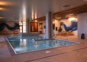 Indoor hotel pool area with turquoise water, a stepped entry and handrail, and wooden lounge chairs along a wall featuring a blue wave mural.