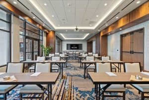Conference room set up for a training or seminar with rows of rectangular tables and white chairs, a projector screen at the far end displaying “WELCOME,” and large windows along the left wall.