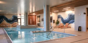 Indoor pool area with a connected hot tub, tiled steps, wooden lounge chairs, and a blue wave mural along the walls.