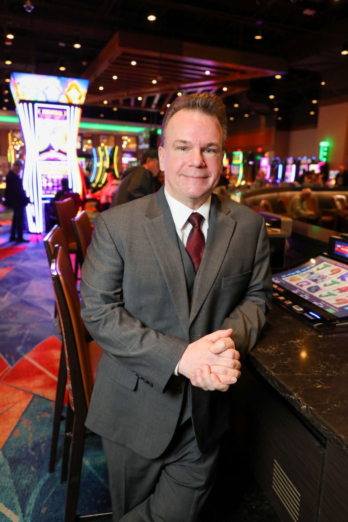 Smiling man in a gray suit and red tie seated at a casino slot-machine area with bright neon lights in the background.