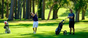 Two men on a sunny golf course: one in mid-swing with a driver, the other standing with arms crossed beside a golf bag on a push cart.