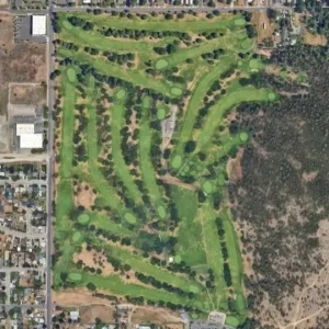 Aerial, top-down view of a winding golf course with multiple greens, fairways, and scattered trees, bordered by residential blocks on the left and native scrub on the right.