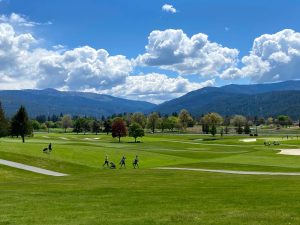 Wide-shot golf course with green fairways, players pushing carts, and a mountain ridge under a bright blue sky with fluffy clouds.