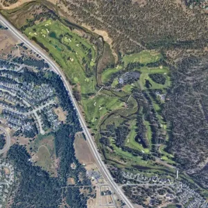 Aerial view of a green golf course winding through trees next to a highway and residential area.