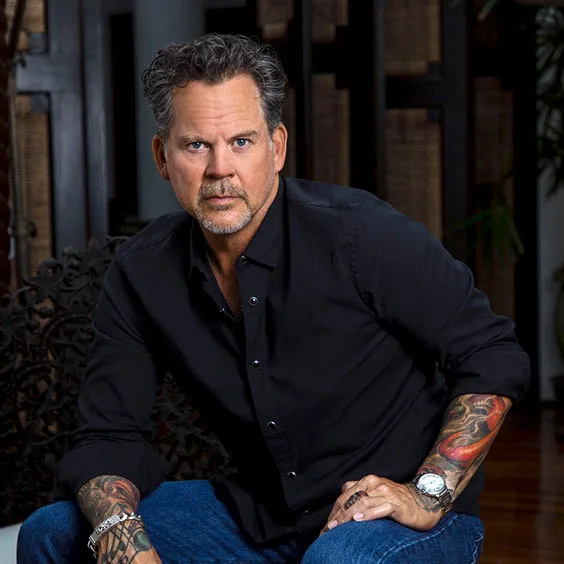 Portrait of a tattooed man in a black button-down shirt seated indoors, looking at the camera.