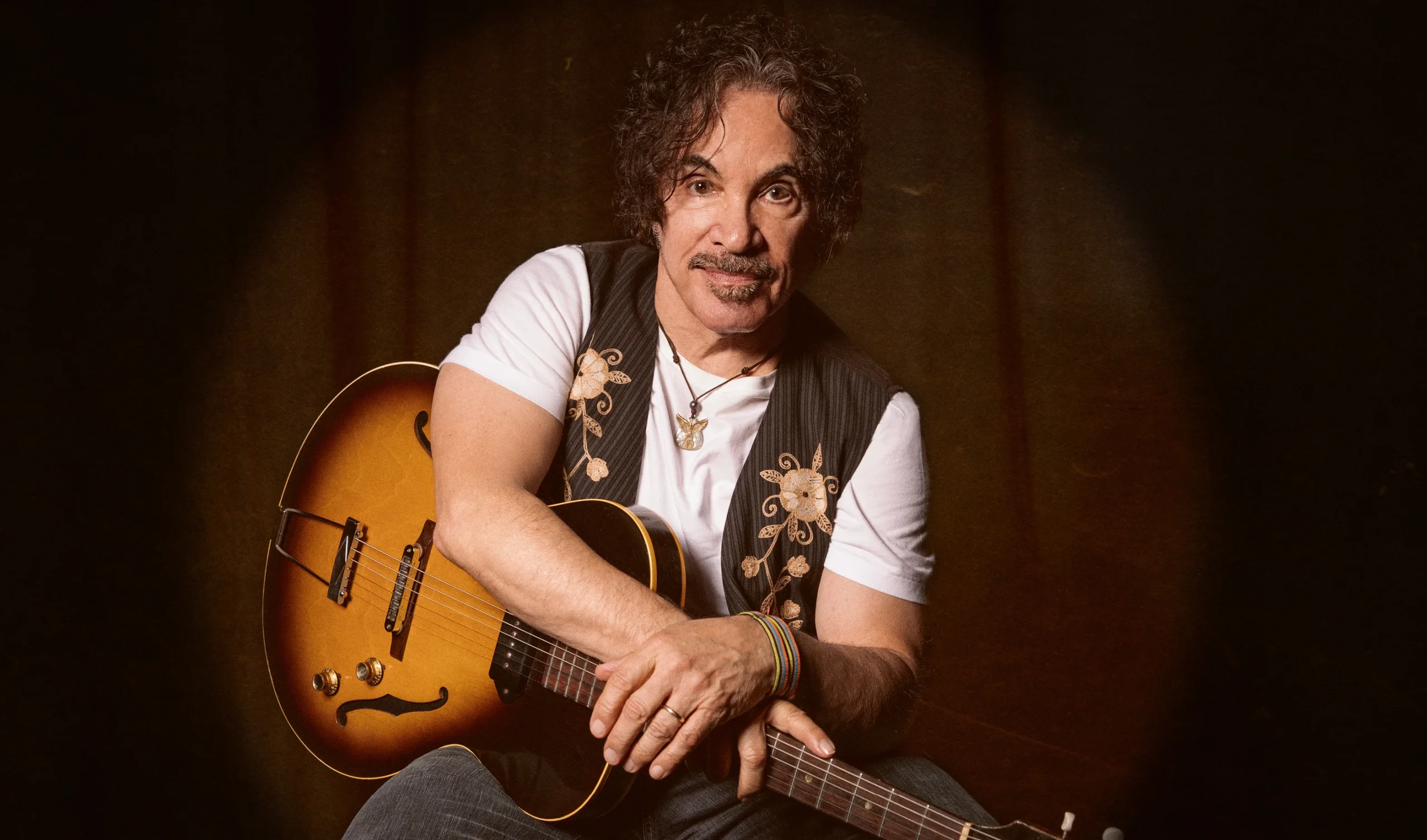 Older man with dark hair and mustache sits, holding a sunburst acoustic guitar on a dark stage backdrop.